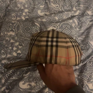 Rutig keps  - Snygg rutig keps i klassiskt beige, svart och rött mönster. Kepsen har justerbar passform baktill och är perfekt för en stilren look. Märkeslogga på baksidan ger en exklusiv touch.