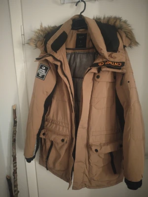 Beige dunjacka med fuskpäls från CMP - Snygg beige dunjacka från CMP med svarta detaljer och avtagbar huva med fuskpäls. Jackan har flera fickor, dragkedja och tryckknappar samt patch på ärmen. Perfekt för kalla dagar och vinteräventyr.