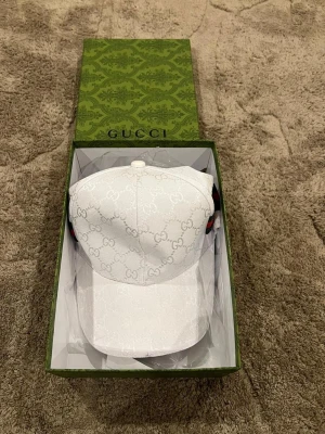 Vit keps med monogram - Säljer en stilren vit keps med det klassiska -monogrammet över hela kepsen. Kepsen har böjd skärm och justerbar rem baktill med röd-gröna detaljer. Kommer i original grön-box. Perfekt för dig som gillar exklusiva accessoarer. Skriv innan köp