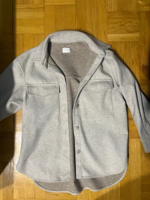 Beige overshirt med fickor - Stilren beige overshirt med klassisk krage och stora bröstfickor. Modellen är rak och har knäppning framtill med fyra knappar. Materialet känns mjukt och har en lätt struktur, perfekt för lager på lager. Passformen är loose och ärmarna är långa.