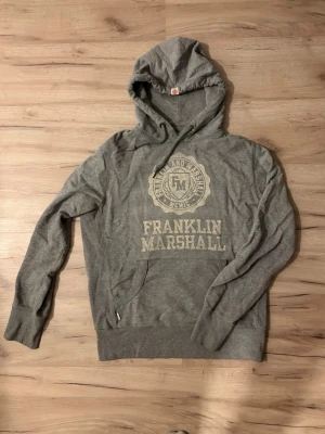 Sällsynt Grå hoodie från Franklin & Marshall - Snygg grå hoodie från Franklin & Marshall med stor logga och text på bröstet. Klassisk känguruficka framtill, dragsko i huvan och ribbade muddar. Perfekt för en avslappnad och sportig stil. Tillverkad i mjukt bomullsmaterial. LIMITED EDITOON SOM EJ SÄLJS MER