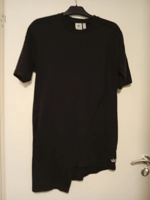 Svart t-shirt klänning från Adidas Originals - En svart t-shirt klänning från Adidas Originals med klassisk rund hals och korta ärmar. T-shirten har en liten Adidas-logga broderad nere vid fållen och är gjord i mjuk bomull. Passar perfekt till en avslappnad streetwear-look.