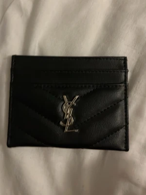 Svart YSL kortfodral i läder - Säljer ett svart kortfodral från Saint Laurent med klassisk YSL-logga i silver framtill. Fodralet är gjort i quiltat läder och har flera kortfack. Perfekt för dig som vill ha en stilren och lyxig accessoar i fickan.