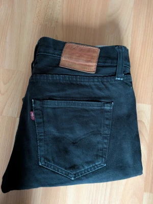 Svarta Levi's 501 jeans W30 L30 - Klassiska svarta Levi's 501 jeans med rak passform och fem fickor. Jeansen har den ikoniska läderpatchen bak i midjan och röd Levi's-tag på bakfickan. Tillverkade i slitstarkt bomullstyg, perfekta för en avslappnad och tidlös stil.