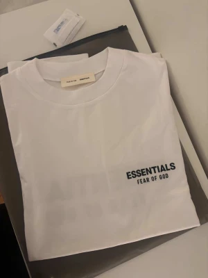 Vit Essentials Fear of God t-shirt - Snygg vit t-shirt från Fear of God Essentials med svart logotyptryck på bröstet. Klassisk rund halsringning och korta ärmar. Tillverkad i mjuk bomull för en clean och stilren look.