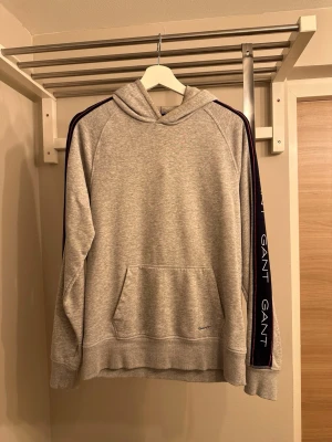 Grå hoodie från GANT med logga - Snygg grå hoodie från GANT med huva och känguruficka. Svarta band med vit GANT-logga längs ärmarna och diskret broderad logga framtill. Tillverkad i mjuk bomullsblandning, perfekt för en avslappnad och trendig look.