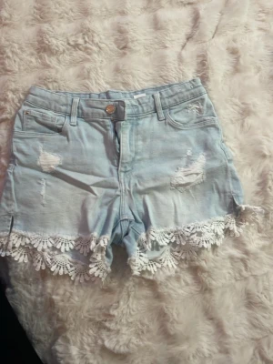 Ljusblå jeansshorts med spetsdetalj - Säljer ett par ljusblå denimshorts med slitna detaljer och vita spetsvolanger längs bensluten. Shortsen har fem fickor, bälteshällor och knappgylf. Perfekta för sommaren och ger en söt, avslappnad look. Storlek 146 men kan användas som xs