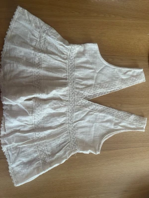 Vit spetsdetaljerad linnetopp från Zara - Söt vit linnetopp från Zara med breda axelband och v-ringad fram- och baksida. Toppen har fina spetsdetaljer och volangkant nedtill. Perfekt för varma dagar och ger en romantisk vibe till din outfit.