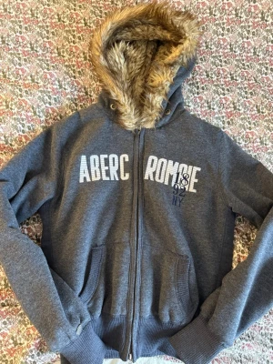 Grå zip-up hoodie från Abercrombie & Fitch med fuskpäls - Mysig grå hoodie från Abercrombie & Fitch med dragkedja, stora fickor och vit logga framtill. Huvan är fodrad med mjuk fuskpäls i beige för extra värme och stil. Perfekt för kalla dagar och chill outfits.
