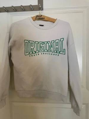 Vit sweatshirt från Gina Tricot - Vit sweatshirt från Gina Tricot med grön text på bröstet. Tröjan har rund halsringning, ribbade muddar vid ärmslut och nederkant samt en avslappnad passform. Perfekt för en casual look.