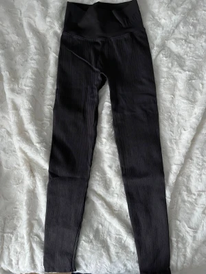 Svarta ribbade leggings med hög midja från Stronger - Snygga gråa leggings från Stronger med ribbad struktur och hög midja. Stretchigt material som sitter tight och formar sig efter kroppen. Sällan använda och i gott skick.
