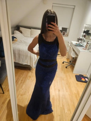 Blå långklänning med spets och paljetter - Elegant blå långklänning med svarta spetsdetaljer och paljetter i midjan och på axelbanden. Klänningen har en öppen rygg och figurnära passform som framhäver siluetten. Perfekt för bal eller festliga tillfällen.