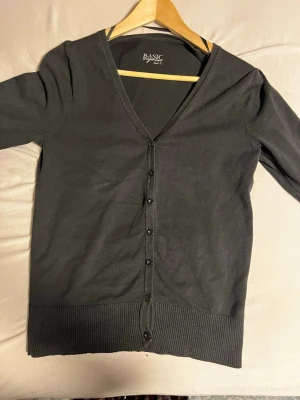 Svart basic kofta med knappar - En enkel svart kofta från BASIC med v-ringning och knäppning framtill. Koftan har långa ärmar och breda ribbade muddar vid ärmslut och nederkant. Perfekt att slänga över en t-shirt eller linne för en clean look.