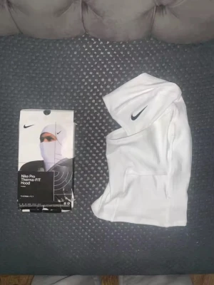 Vit Nike Pro Shiesty Therma-FIT Hood - Nike Pro Shiesty Therma-FIT Hood i vitt, tillverkad i mjukt och stretchigt material. Huva med svart Nike-logga på sidan, designad för att ge värme och skydd. Perfekt för träning utomhus eller kalla dagar.