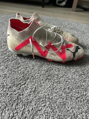 Beige och röda Puma fotbollsskor - Säljer ett par beige fotbollsskor från Puma med röda och svarta detaljer. Skorna har högre strumpkrage, snörning och är tillverkade i syntetmaterial för bra passform på planen. Perfekta för dig som vill sticka ut med stilrena och sportiga skor.obs ingen påse med