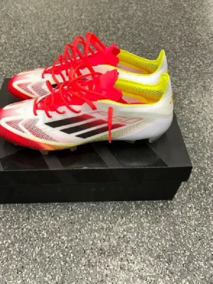 Adidas vita och röda fotbollsskor - Säljer ett par Adidas fotbollsskor i vitt med röda och gula detaljer samt svarta ränder på sidan. Skorna har snörning och är tillverkade i syntetmaterial för lätt känsla på planen. Perfekta för dig som vill sticka ut med färg och stil på matchen. Storleken passar också 40/5