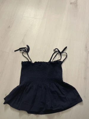 Marinblå linnetopp med knytband - Söt marinblå linnetopp med smockad överdel och volang nertill. Axelbanden är tunna och knyts upptill för en justerbar passform.