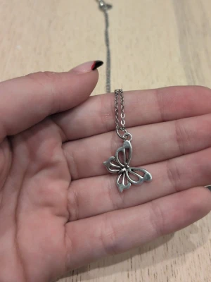Silverfärgat halsband med fjärilshänge i rostfritt stål  - Snyggt halsband i silverfärg med ett öppet, stilrent fjärilshänge. Kedjan är tunn och smidig, och fjärilen har en modern, minimalistisk design. Perfekt för dig som gillar enkla men unika accessoarer. Materialet ser ut att vara metall.
