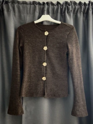 Brun stickad kofta med blommiga knappar - Mysig brun stickad kofta med långa ärmar och unika blommiga knappar framtill. Koftan har en rak passform och är tillverkad i ett mjukt, ribbat material som känns skönt mot huden. Perfekt för lager på lager och en stilren vardagslook.