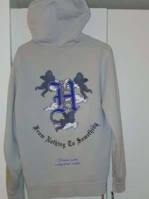 Grå hoodie från Hoodrich med tryck - Snygg grå hoodie från Hoodrich med stor svart text på bröstet och coola änglamotiv både fram och bak. På ryggen står det 'From Nothing To Something' och ett stort H i blått. Klassisk känguruficka och huva. Perfekt för en streetwear-look.