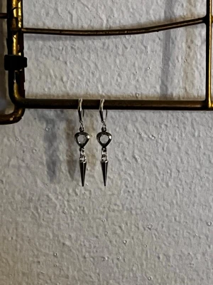 Silvriga örhängen med spetsig hänge - Snygga örhängen i silverfärgad metall med hjärtformad detalj och hängande spetsig kon. Perfekta för dig som gillar en edgy och trendig stil. Lätta att bära och passar till många olika looks.