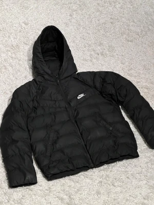 Svart pufferjacka från Nike jr - Svart pufferjacka från Nike med huva och vit logga på bröstet. Jackan har dragkedja framtill, två sidofickor med knappstängning och quiltad design. Perfekt för kalla dagar och har en clean, sportig look som funkar till streetwear.