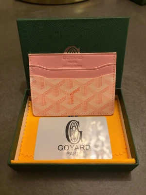 Rosa kortfodral från G - Supersnyggt rosa kortfodral från G med klassiskt mönster och flera kortfack. Tillverkat i skinn med Goyards ikoniska tryck i ljusrosa och vitt. Perfekt för dig som vill ha en stilren och lyxig accessoar i fickan.