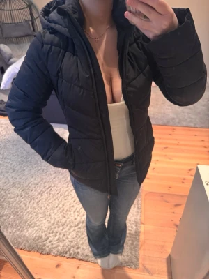 Svart pufferjacka med huva från Zara  - Säljer en svart pufferjacka med huva och dragkedja framtill. Jackan har en quiltad design och normal passform, perfekt för kyliga dagar. Enkel och stilren look som passar till det mesta.