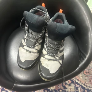 Adidas Terrex grå kängor med snörning - Adidas Terrex kängor i grått och svart med orange detaljer. Skorna har snörning, robust sula och är tillverkade i syntetmaterial och textil. Perfekta för utomhusaktiviteter och har en sportig design med tydliga logotyper på sidan.