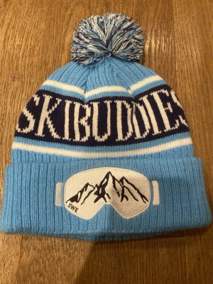 Blå SKIBUDDIES mössa med tofs - Säljer en ljusblå ribbstickad mössa med vit och mörkblå texten 'SKIBUDDIES' runtom. Mössan har en stor tofs på toppen och ett broderat märke med skidglasögon och bergsmotiv framtill. Perfekt för dig som gillar vinter och skidåkning.