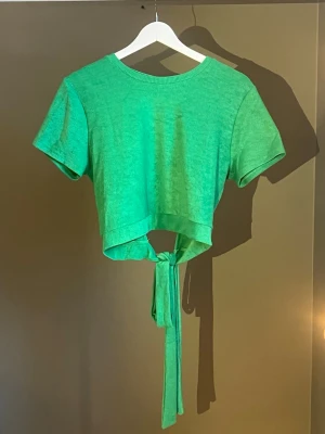 Grön croppad topp med knyt - Trendig grön croppad topp med korta ärmar och snygg öppen rygg. Toppen har breda band som knyts i midjan för en cool detalj. Perfekt för dig som vill sticka ut med en färgstark och unik look. Den är i storlek XL men passar även L. Köpt från Gina Tricot, använts en gång💚🍭