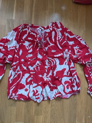 Rödvit blus med mönster från H&M - Säljer en luftig blus från H&M i storlek L med röd och vit abstrakt mönster. Blusen har knappar framtill, långa ärmar med resår vid ärmslut och en avslappnad passform. Perfekt för dig som gillar färgstarka plagg och vill sticka ut.