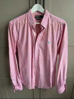 Rosa rutig skjorta från Polo Ralph Lauren - Säljer en rosa och vit rutig skjorta från Polo Ralph Lauren i slim fit. Skjortan har långa ärmar, klassisk krage och knappar framtill. Tillverkad i mjuk bomull och har den ikoniska broderade loggan på bröstet.