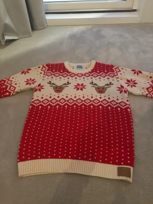 Röd jultröja med renar och mönster - Julig stickad tröja i rött och vitt med renar och klassiskt vintermönster. Rund halsringning och långa ärmar. Perfekt för mysiga vinterdagar och julfirande.