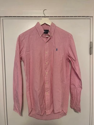 Rosa rutig skjorta från Ralph Lauren - Säljer en rosa och vit rutig skjorta från Ralph Lauren i modellen Custom Fit. Skjortan har lång ärm, klassisk krage, knappar framtill och den ikoniska blå loggan broderad på bröstet. Perfekt för dig som gillar stilrena och tidlösa plagg.