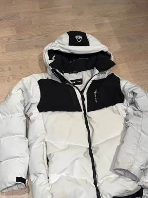 Vit och svart pufferjacka från sailRacing - Snygg pufferjacka från sailRacing i vit och svart med huva och dragkedja framtill. Jackan har quiltad design, hög krage och två sidofickor. Perfekt för kalla dagar och ger en sportig vibe. Säljer jackan nu eftersom jag inte har behov av den längre, eftersom jag vuxit ur den. 