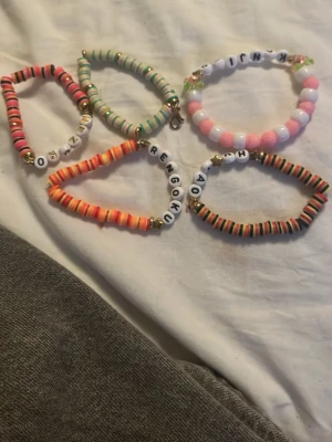 Färgglada pärlarmband med bokstäver - Säljer flera handgjorda armband med färgglada plastpärlor och bokstavspärlor. Armbanden har olika färgkombinationer som rosa, orange, grön och svart samt detaljer i guld. Några armband har namn eller ord med bokstäver. Perfekt accessoar för en personlig touch!