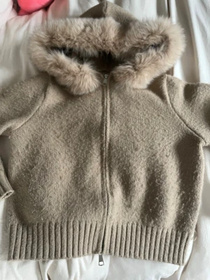 Beige teddyjacka med fuskpäls - Mysig beige teddyjacka med huva och fluffig fuskpäls runt kanten. Jackan har dragkedja framtill och bred ribbad mudd nedtill. Perfekt för kyliga dagar när du vill vara både varm och stylish.