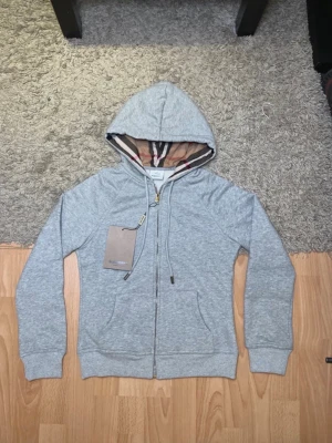 Grå zip hoodie från Burberry - Snygg grå zip hoodie från Burberry med klassiskt rutigt foder i huvan och broderad logga på bröstet. Tillverkad i mjuk bomullsblandning, har dragsko i huvan och två fickor framtill. Perfekt för en chill och stilren look.