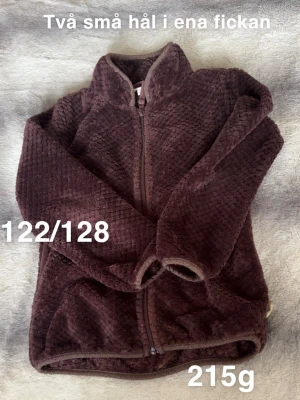  fleecekofta med dragkedja - Supermjuk och fluffig fleecekofta med hel dragkedja och hög krage. Koftan har lång ärm och en mysig struktur som håller dig varm. Perfekt för lager-på-lager och chill dagar. Storlek 122/128.🌸 Två små hål i ena fickan. Storlek 122/128. Brun/lila i färg