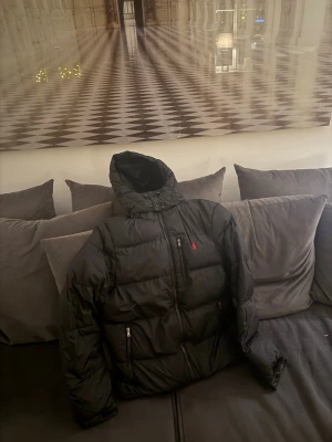 Svart dunjacka från Polo Ralph Lauren - Säljer en svart dunjacka från Polo Ralph Lauren med huva och dragkedja framtill. Jackan har två framfickor med dragkedja och den klassiska röda Polo-loggan på bröstet. Perfekt för kalla vinterdagar och riktigt skön att bära.