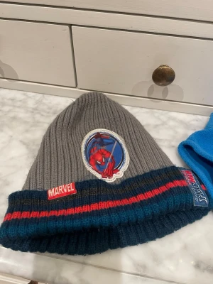 Marvel Spiderman mössa grå/blå - One size. För barn ca 5-7 år. Stickad mössa i grått med blå och röda ränder från Marvel. Framtill finns ett broderat Spiderman-märke och Marvel-logga. Mössan har ribbstickad kant och är perfekt för dig som gillar superhjältar och vill ha en cool accessoar till vintern.