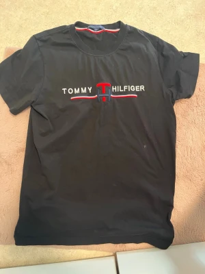 Svart t-shirt från Tommy Hilfiger - Svart t-shirt från Tommy Hilfiger med broderad logga i vitt, rött och blått på bröstet. Klassisk rund halsringning och korta ärmar. Tillverkad i mjuk bomull för en bekväm känsla.