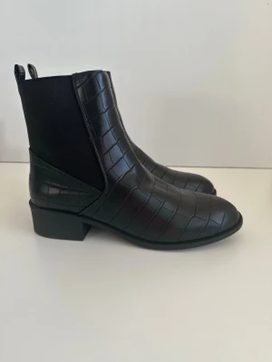 Svarta Chelsea boots - helt NYA - Ett par HELT NYA svarta boots / ankelstövlar / chelseaboots från Miss Selfridge i storlek 40. Superfint veganskt konstläder med krokodilprägling. Praktisk resår på sidorna och bekväm låg klack. Lika snygg till byxor som till klänning.   Hämtas i Stockholm eller skickas mot porto. 