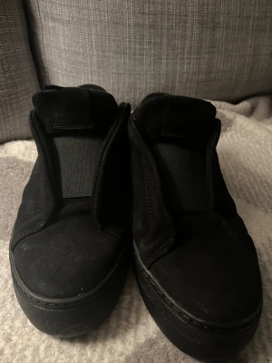 Svarta sneakers från Axel Arigato - Säljer ett par helsvarta sneakers från Axel Arigato i mocka med minimalistisk design. Skorna har rund tå, platt sula och diskret logga på sidan. Passar perfekt till streetwear och har en clean look utan snörning. Nypris 2300kr