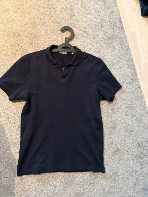 Mörkblå pikétröja från Bläck - En stilren mörkblå pikétröja från Bläck i klassisk modell med krage och korta ärmar. Tillverkad i mjuk bomull som känns skön mot huden. Perfekt för en clean och enkel look.