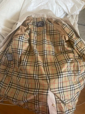 Beige trenchcoat från Burberry - Endast använd ett par gånger, men tyvärr för stor. Rejält tyg. Klassisk beige trenchcoat från Burberry med det ikoniska rutiga fodret. Jackan har  knappar och raka fickor med knapp framtill. Perfekt för dig som vill ha en tidlös och stilren look. Som ni ser på en av bilderna: Etiketten i fickan står det ”52 short”. Den är äkta. Jag har fått den, men flera har bekräftat att den är äkta. Detta var innan de ändrade sitt namn till Burberry. Så det kan va vintage men är osäker. 