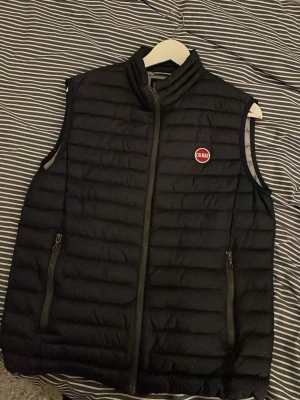 Svart dunväst från Colmar - Snygg svart dunväst från Colmar med quiltad design och hög krage. Västen har två dragkedjeförsedda fickor framtill och Colmar-logga på bröstet. Perfekt att slänga över en hoodie eller tröja för extra värme och stil.