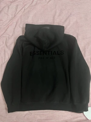 Svart Essentials Fear of God hoodie - Svart hoodie från Essentials Fear of God med stor huva och känguruficka. Tryck med 'ESSENTIALS' och 'FEAR OF GOD' på ryggen samt mindre logga på bröstet. Tillverkad i mjukt bomullsmaterial, perfekt för en avslappnad streetwear-look.