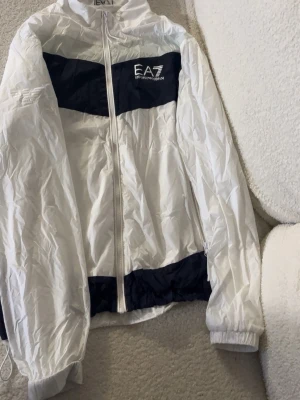 Vit och marinblå EA7 vindjacka - Snygg vindjacka från EA7 Emporio Armani i vitt med marinblåa detaljer. Jackan har dragkedja framtill, hög krage och elastiska muddar. EA7-logga på bröstet och präglad logga på ärmen. Tillverkad i lätt, prassligt material som passar perfekt till streetwear.